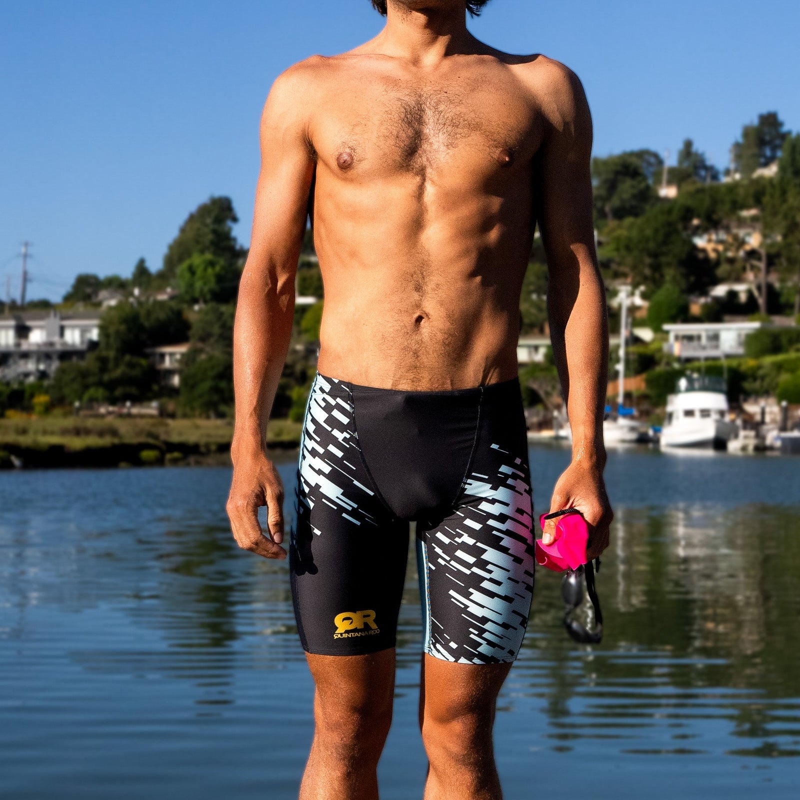 Man wearing black and white swim shorts standing by a waterfront with boats and houses in the background.