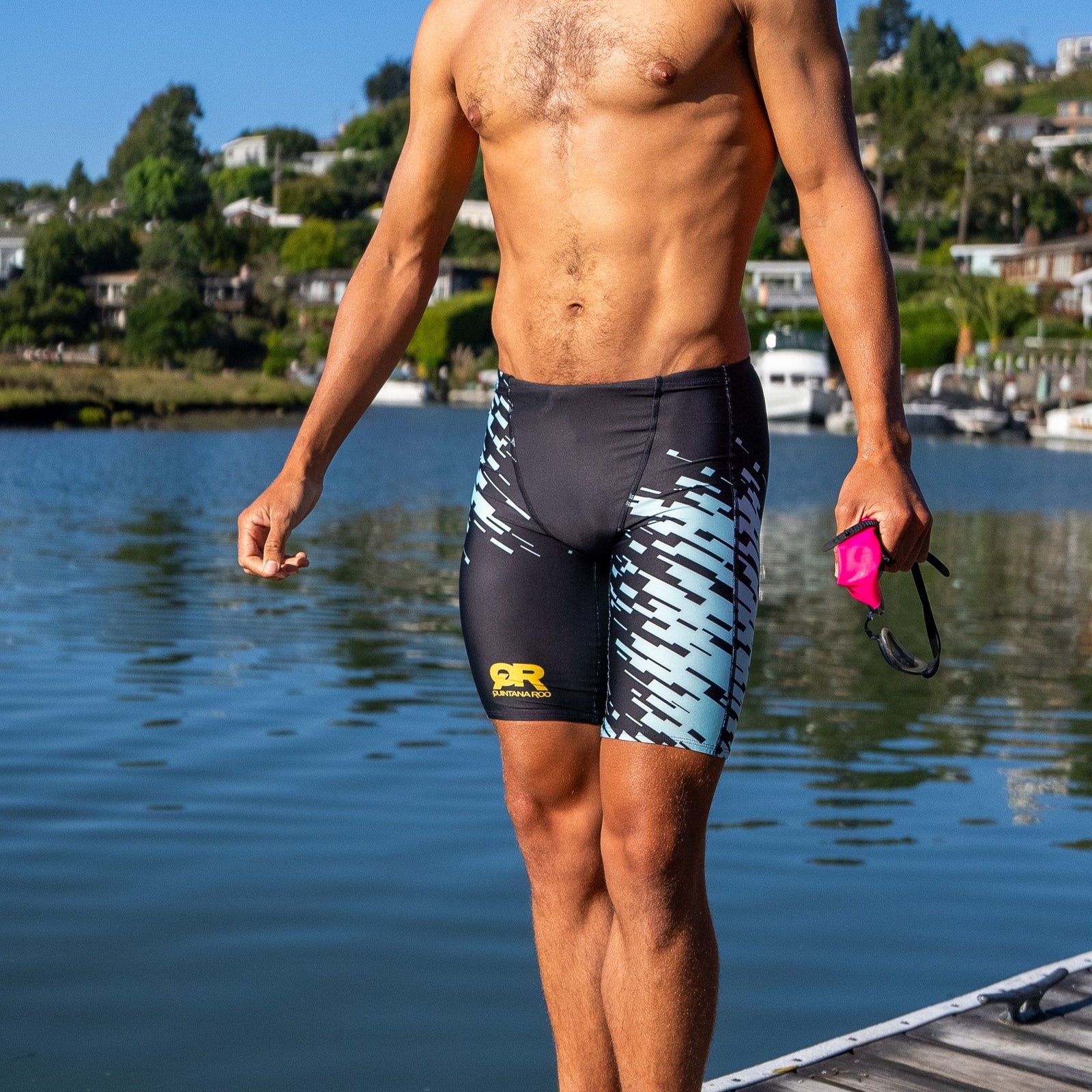 Man standing on a dock by a lake wearing swim shorts with a QR logo.