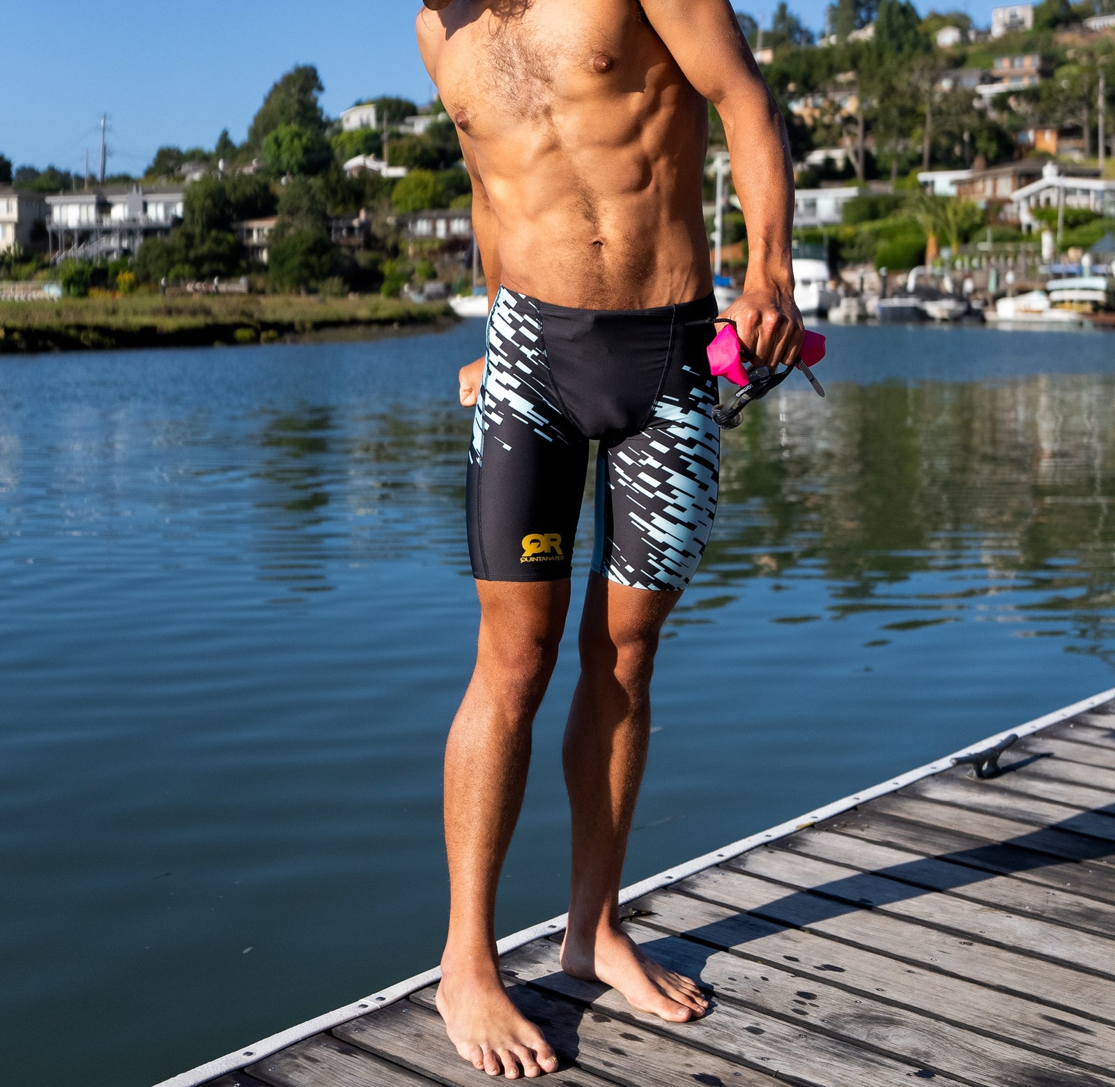 Man in swim shorts standing on a dock by a body of water with houses in the background