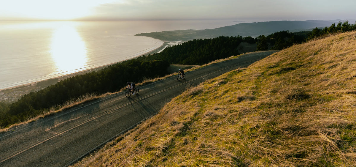 Two cyclists ride along a winding coastal road at sunset, with ocean water, shoreline, forested hills, and a sunlit grassy slope in the foreground.