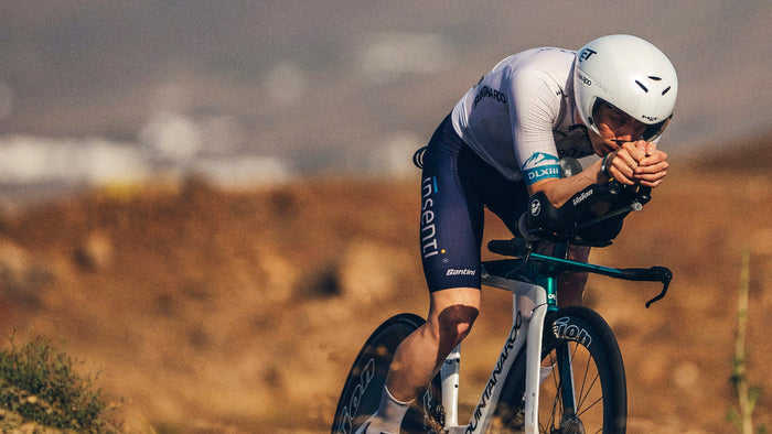 A cyclist in a white and navy team jersey leans forward on a time-trial bicycle in an outdoor desert setting with dry brown hills in the background.
