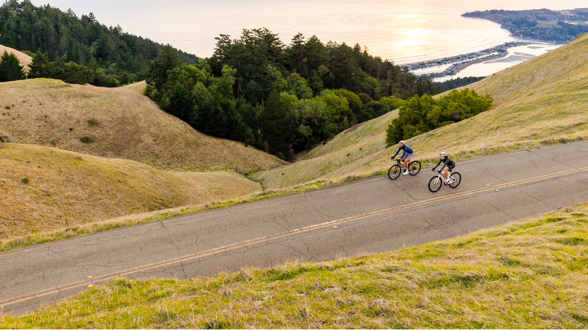 Two cyclists ride along a winding paved road on grassy hills overlooking a coastal bay at sunset, with trees and distant shoreline in the background.
