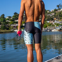 Person wearing swim trunks by a waterfront with houses and boats in the background