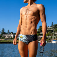 Man wearing swim trunks standing by a waterfront with boats and buildings in the background