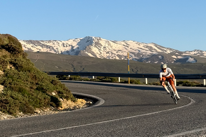 A cyclist in a white and orange cycling jersey rides a road bike along a winding asphalt road with a backdrop of snow-capped mountains and a clear blue sky.